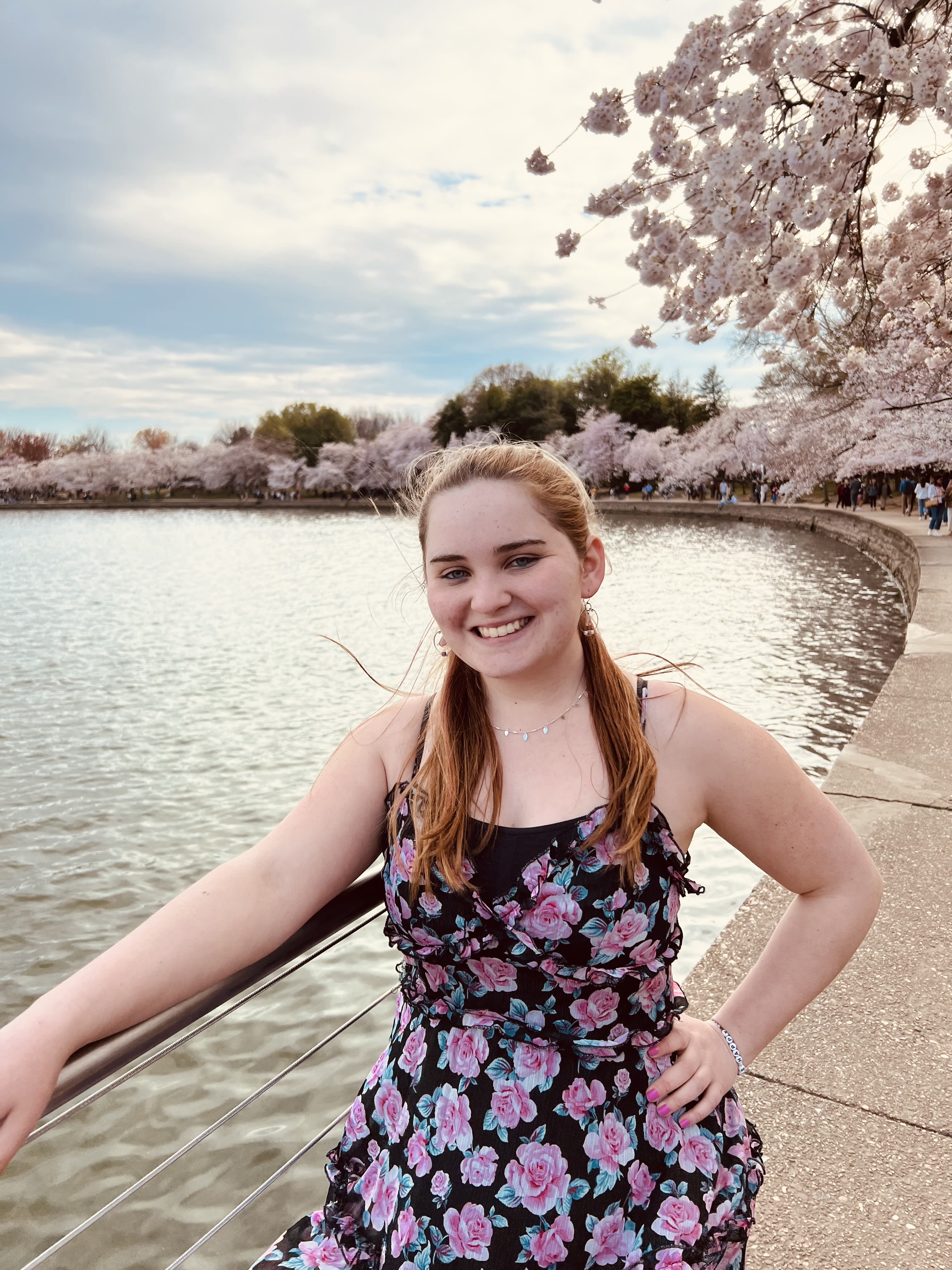 Lilly Shaw, wearing a black dress with floral pink print, posing and smiling beside pink cherry trees and a body of water.