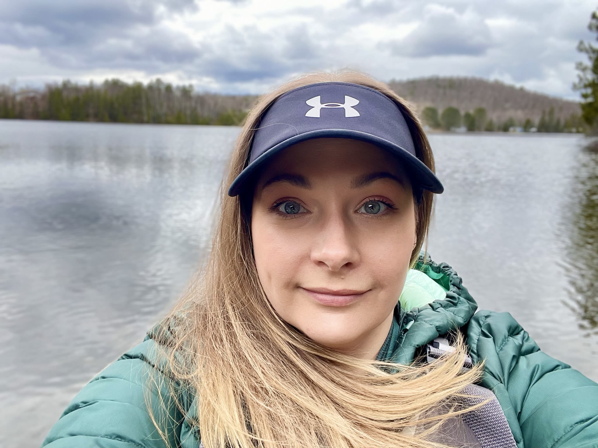 • A selfie of Jessica O’Reilly from the shoulders up, standing in front of a lake wearing a green jacket and blue Under Armor visor.