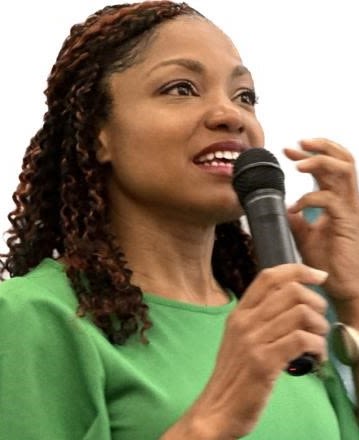 T. Nicole Tucker-Smith, a Black woman in a green dress speaking into a microphone and looking hopeful.